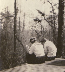 Two Bryn Mawr students in white athletic clothing of the early 20th century sitting on a porch, their backs to the viewer. A forest of fir trees is visible in the middle-distance.